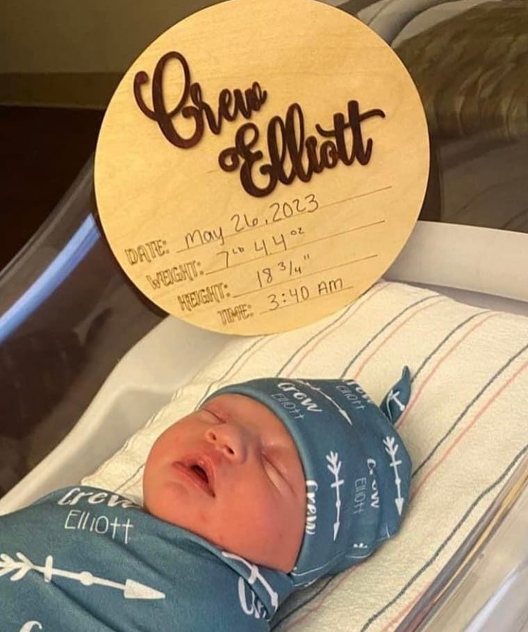 A Newborn Baby Named Crew Elliott Swaddled In A Personalized Blue Blanket, With A Bamboo Birth Announcement Tag Beside Him Displaying His Name, Birth Date, Weight, Height, And Time.