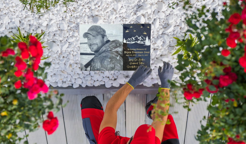 Person Arranging A Laser-Engraved Home Garden Memorial Plaque With A Portrait And Inscription, Surrounded By White Stones And Red Roses.