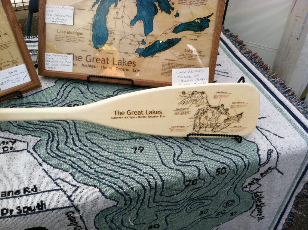 A wooden canoe paddle engraved with a map of the Great Lakes, displayed on a table with other framed maps of the region.