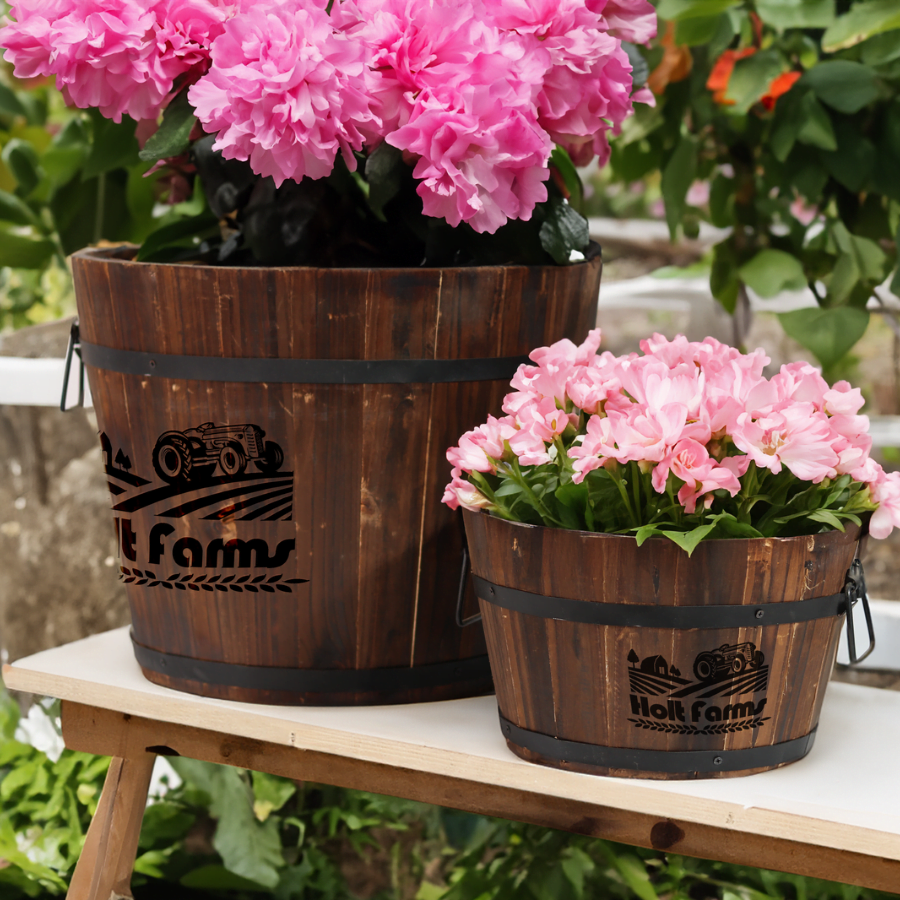 Two wooden planters engraved with "Holt Farms" logo, filled with bright pink flowers, placed outdoors in a garden setting.